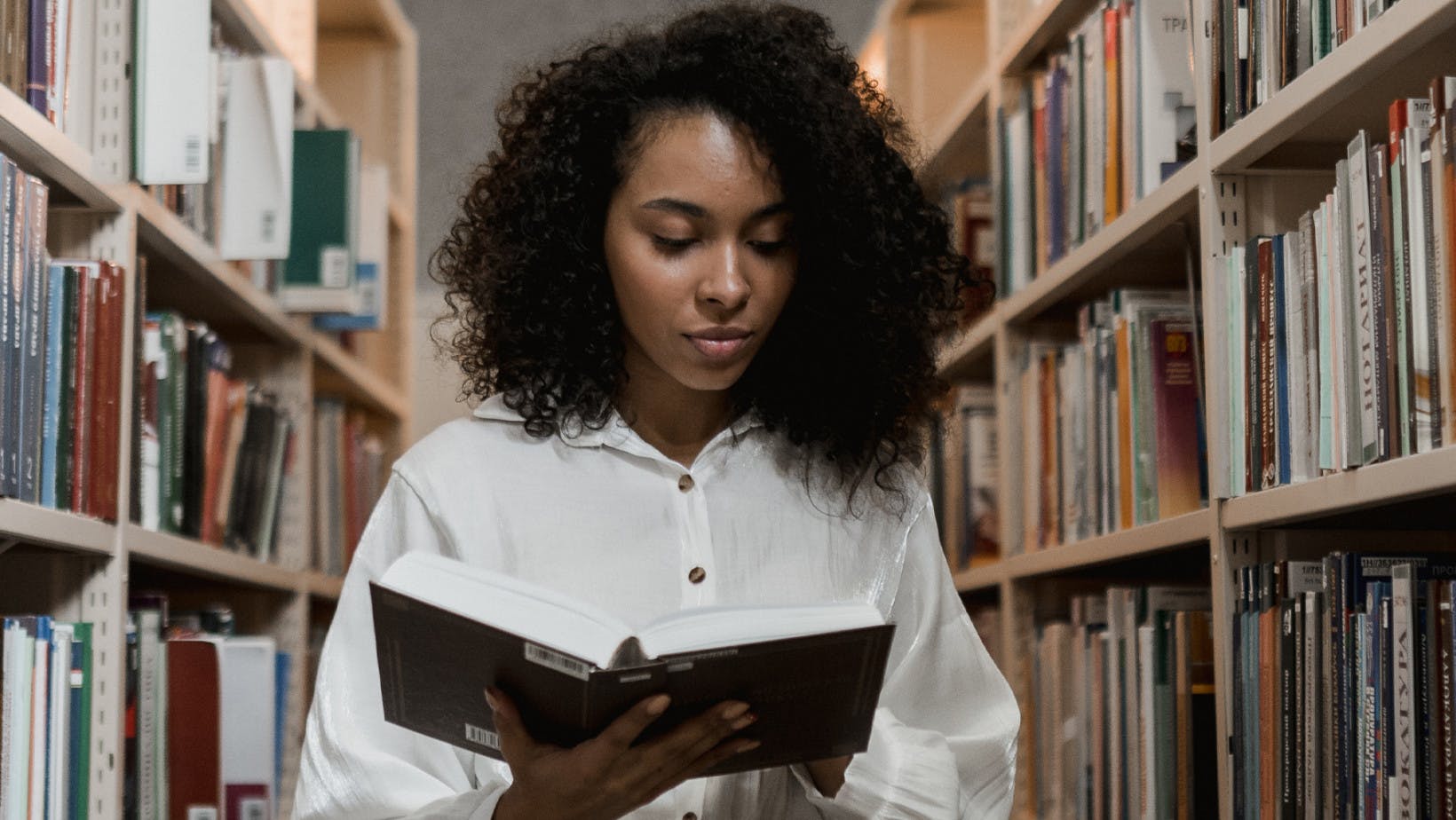 Black woman reading a literature book in a library aisle