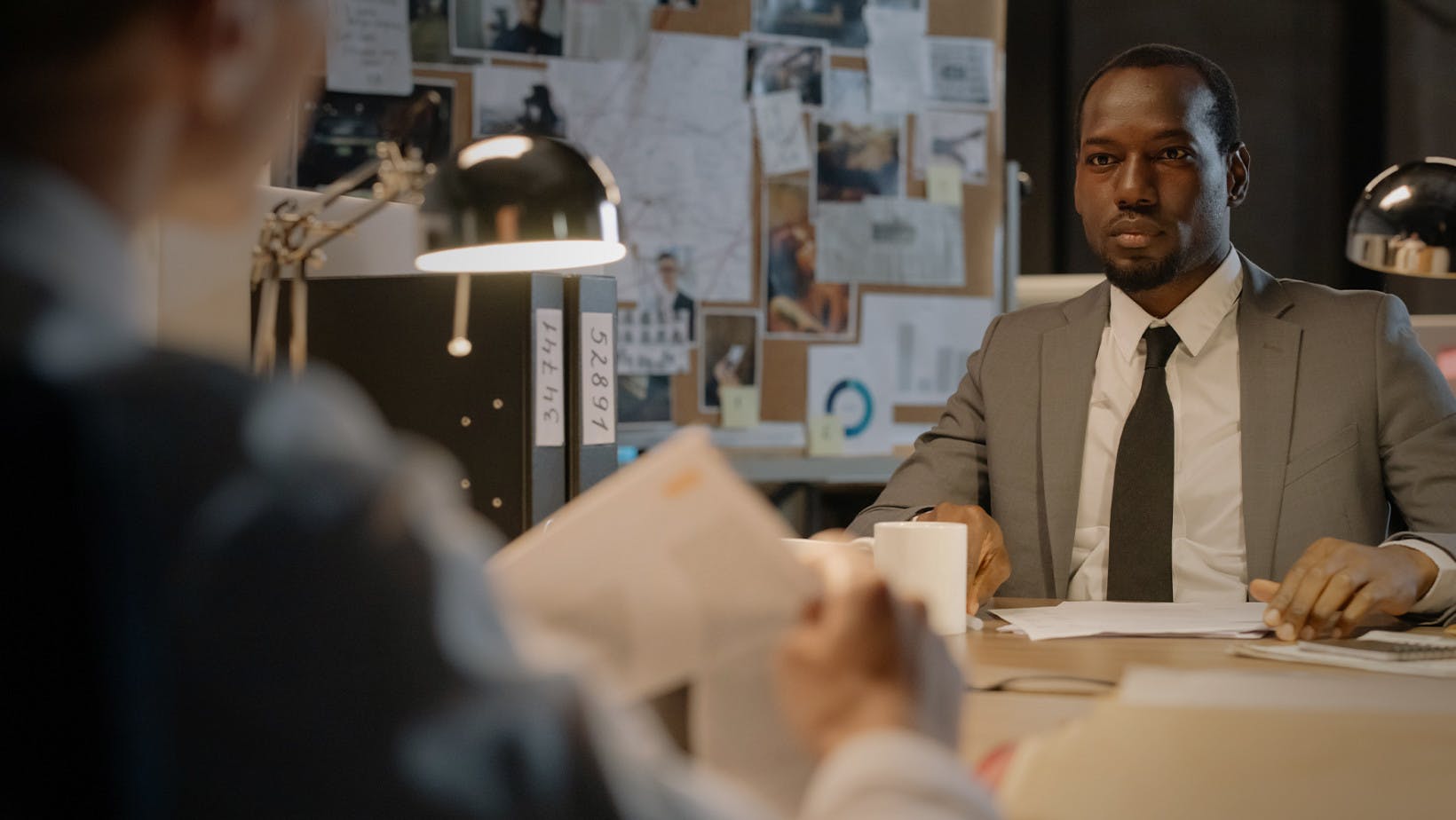 A black man who is a Criminal Psychologist sits at a desk with a colleague blurred in the foreground