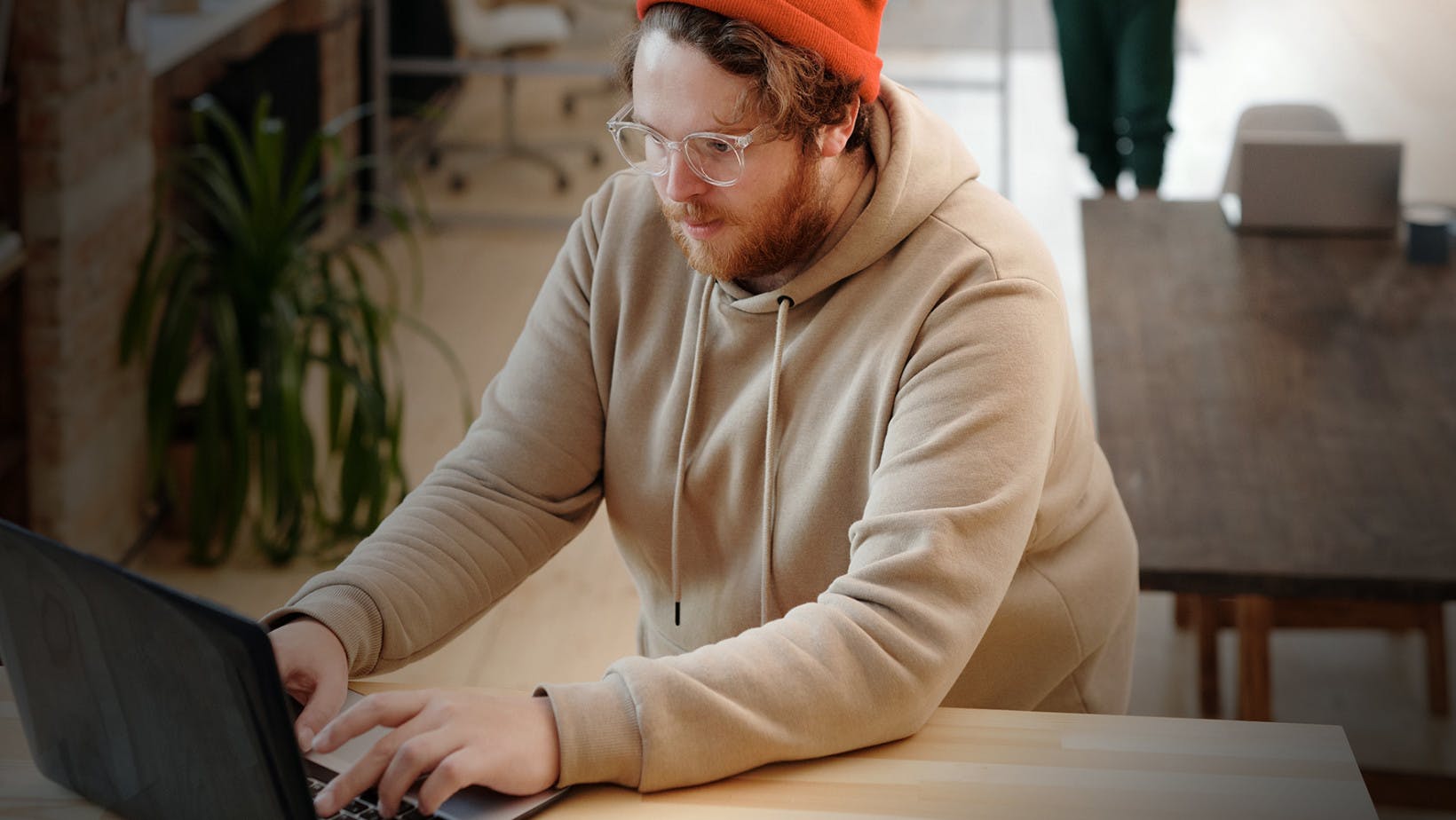 Male student with a hoodie and glasses on is researching scholarships on a laptop